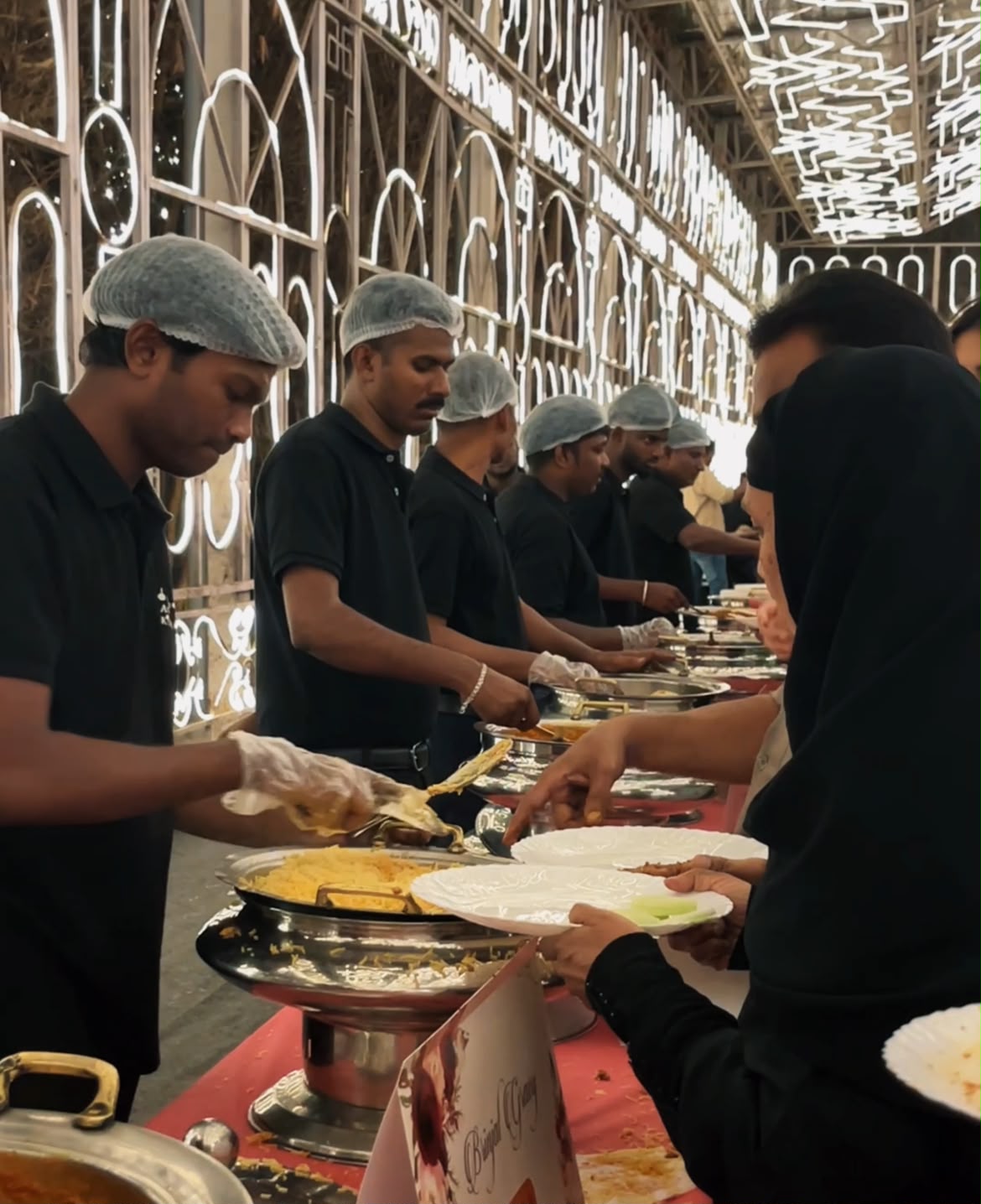 Uniformed catering team serving biryani at a lit wedding venue
